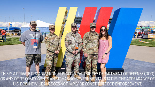 Group of U.S. military service members and a NASCAR representative posing in front of a large NASCAR logo at the track. Disclaimer text notes no affiliation or endorsement by the Department of Defense.
