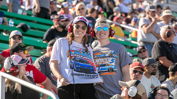 Two NASCAR fans in the grandstands smiling while holding a scanner radio, wearing race team shirts and headsets with the crowd cheering in the background