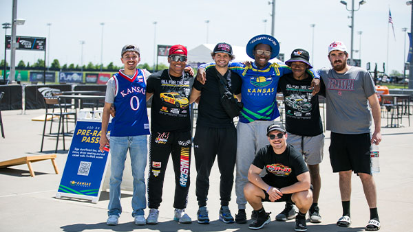 Group of college students and friends enjoying a day at the track, wearing Kansas apparel and NASCAR gear, smiling together for a group photo.