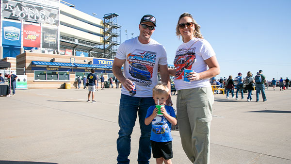 Teacher and family at the Kansas Speedway, standing outside ticket gates. Parents wearing race-themed shirts with their young child holding a drink in the center.