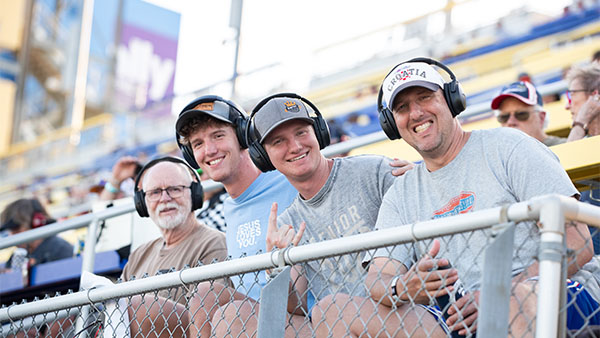 Four NASCAR fans sitting in the grandstands wearing headsets, smiling and enjoying the race from their reserved seats on a sunny day.