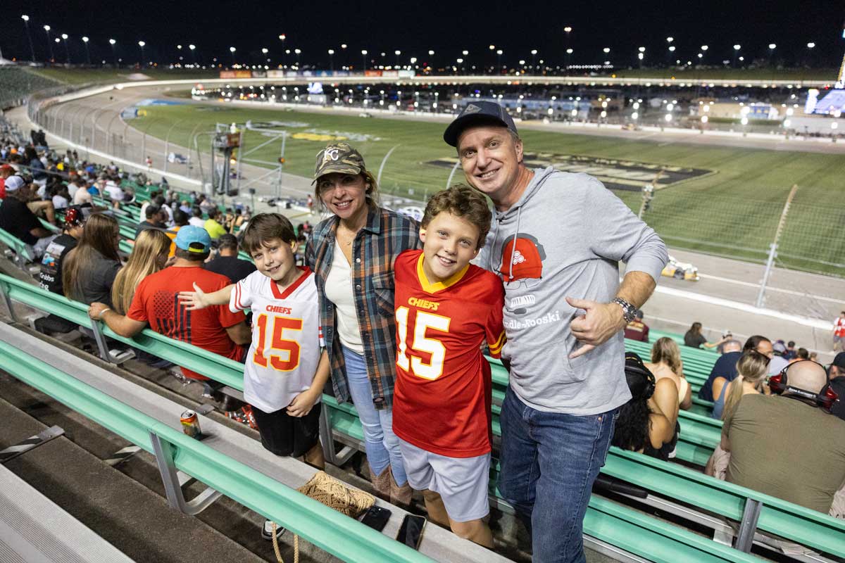 A family of four poses and smiles in the grandstands at a nighttime motorsports event. Two children wearing red and white Kansas City Chiefs jerseys stand in front of two adults, with the illuminated race track and infield visible behind them. Spectators fill the surrounding seats, and stadium lights brighten the venue under the night sky.